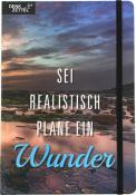 Notizbuch mit einem Landschaftsbild auf dem Cover. Text: 'SEI REALISTISCH PLANE EIN Wunder'. Farben: Blau, Grau, Orange.