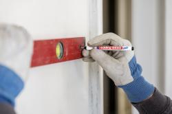 Hand mit grauen Arbeitshandschuhen, die einen roten Wasserwaage hält. Ein schwarzer Marker wird an die Wand gehalten. Hintergrund ist hell.