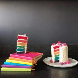 Bunter Regenbogenkuchen mit mehreren Schichten. Neben dem Kuchen liegen farbige Notizbücher gestapelt.