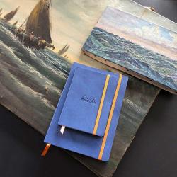 Zwei blaue Notizbücher mit orangefarbenen Gummibändern. Im Hintergrund ein Gemälde mit Segelbooten auf dem Wasser.