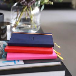 Drei Notizbücher in verschiedenen Farben: blau, rot und pink. Sie liegen auf einem Tisch neben einer Vase mit Blumen.