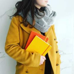 Frau mit braunem Mantel und grauem Schal. Sie hält drei Notizbücher in orange, gelb und rot.