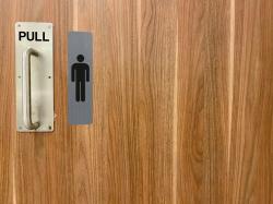 Holztür mit einem silbernen Türgriff. Zeichen für Männer-Toilette daneben.