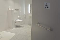 Barrierefreies Badezimmer mit Toilette und Waschbecken. Helle Fliesen und Haltegriffe sind vorhanden.