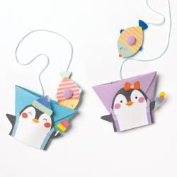 Bastelbox mit bunten Papierfiguren. Enthält Pinguine, Fische, ein Tier mit Pullover und Bastelmaterialien. Farbenfroh und kinderfreundlich.
