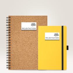Zwei Notizbücher. Ein großes, braunes, spiralförmiges Notizbuch aus Kork. Ein kleines, gelbes Notizbuch mit Gummiband. Beide mit dem Aufdruck 'PROJEKTNOTIZEN'.