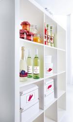 Regal mit verschiedenen Flaschen und Gläsern. Oben Marmelade in Gläsern mit roten Deckeln. Unten Flaschen mit Wein und Sekt. Weiße Boxen mit roten Symbolen.