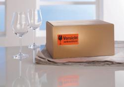 Rote Warnetiketten mit dem Text 'Vorsicht zerbrechlich!'. Verpackung zeigt eine gelbe Box mit Etiketten für den Paketversand.