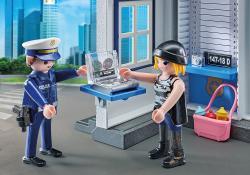 Playmobil-Figuren: Polizist übergibt ein Gerät an eine Person mit Hut. Hintergrund mit modernen Gebäuden.