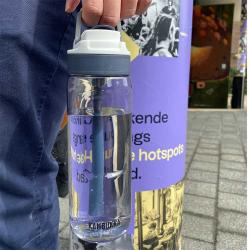 Transparente Wasserflasche mit blauer Kappe und Strohhalm. Flasche steht auf einem Gehweg neben einem Plakat.