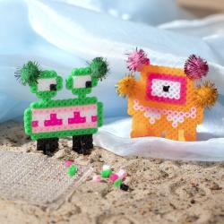 Mini Bastelset mit Bügelperlen. Zwei bunte Monsterfiguren. Eine Figur grün mit rosa Mund, die andere orange mit bunten Pompons.