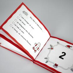 Offenes Weihnachtskalenderbuch mit roten Rahmen, weißen Seiten und kleinen Weihnachtsmotiven. Auf der linken Seite sind Text und eine kleine Schneeflocke, auf der rechten Seite eine große Zahl zwei mit Weihnachtsdeko.