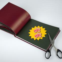 Offenes Buch mit dunklem Hintergrund. Zwei kleine Papierfiguren mit roten Umrissen liegen auf einer gelben Sprechblase.