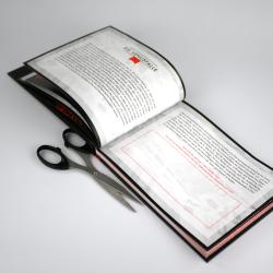 Offenes Buch mit schwarzen Seiten und roten Akzenten. Darin sind Textpassagen sichtbar, das Buch liegt auf einer weißen Oberfläche.