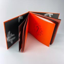 Aufklappbares Buch mit orangefarbenem Einband und schwarzen Innenseiten. Die Innenseiten zeigen weiße Zweige und eine Seite mit einem schwarzen Umriss eines Zuckerstangen-Symbols.