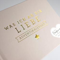 Helles Kartenmaterial mit goldener Prägung. Der Text lautet Was ich an dir liebe Adventskalender mit goldener Sternverzierung.