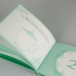 Offenes Buch mit grünen Rahmen und Blättern. Seite zeigt einen Text und eine einfache Illustration einer Tasse mit Dampf.