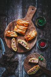 Holzbrett mit mehreren Stückchen gefüllter Pastete. Nebenan kleine Gläser mit grüner und roter Flüssigkeit, dunkler Holzuntergrund.