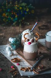 Zwei Gläser mit Schokoladenpudding und Sahne, Dekoration mit Schokoladekugeln und Zuckerstangen. Weihnachtsdeko mit Tannenzweigen, weißen Kugeln und Notenblatt im Hintergrund.