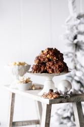 Schokoladen-Haselnuss-Riegel auf einem weißen Kuchenständer, daneben weiße Weihnachtskugeln und Zimtstangen. Im Hintergrund eine verschneite Tannenbaum-Ästhetik.