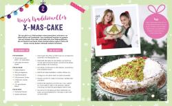 Zwei junge Mädchen präsentieren eine festlich dekorierte Torte mit weihnachtlichem Motiv. Im Hintergrund ein Weihnachtsbaum mit grünen Zweigen und roten Kugeln.