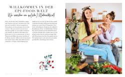 Zwei junge Frauen sitzen auf einer Küchenarbeitsplatte. Eine hält ein Smartphone, beide lächeln freundlich. Im Vordergrund sind frische Kräuter und Gemüse sichtbar.