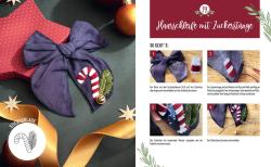 Deko-Element mit einer violetten Stoffschleife, einem kleinen Zuckerstangen-Applikation mit roten und weißen Streifen, einer grünen Zweig- und einer goldfarbenen Glocke. Im Hintergrund eine Geschenkbox mit rotem Deckel und weißen Punkten, sowie goldene und weiße Sternchen.