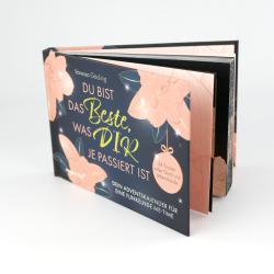 Aufklappbares Adventskalender-Booklet mit rosa, dunkelblau und goldenen Akzenten. Vorderseite mit Blumenmuster und Text, innen Seiten mit weiteren Designs und Text.