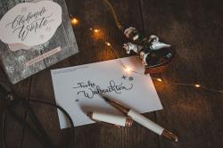 Weißes Briefpapier mit handgeschriebener Weihnachtsbotschaft Frohe Weihnachten. Dekorative Schneeflocken und kleine Schneemänner. Ein Kugelschreiber liegt auf dem Papier, daneben eine Adventskalenderbox mit dem Titel Gluhende Worte.