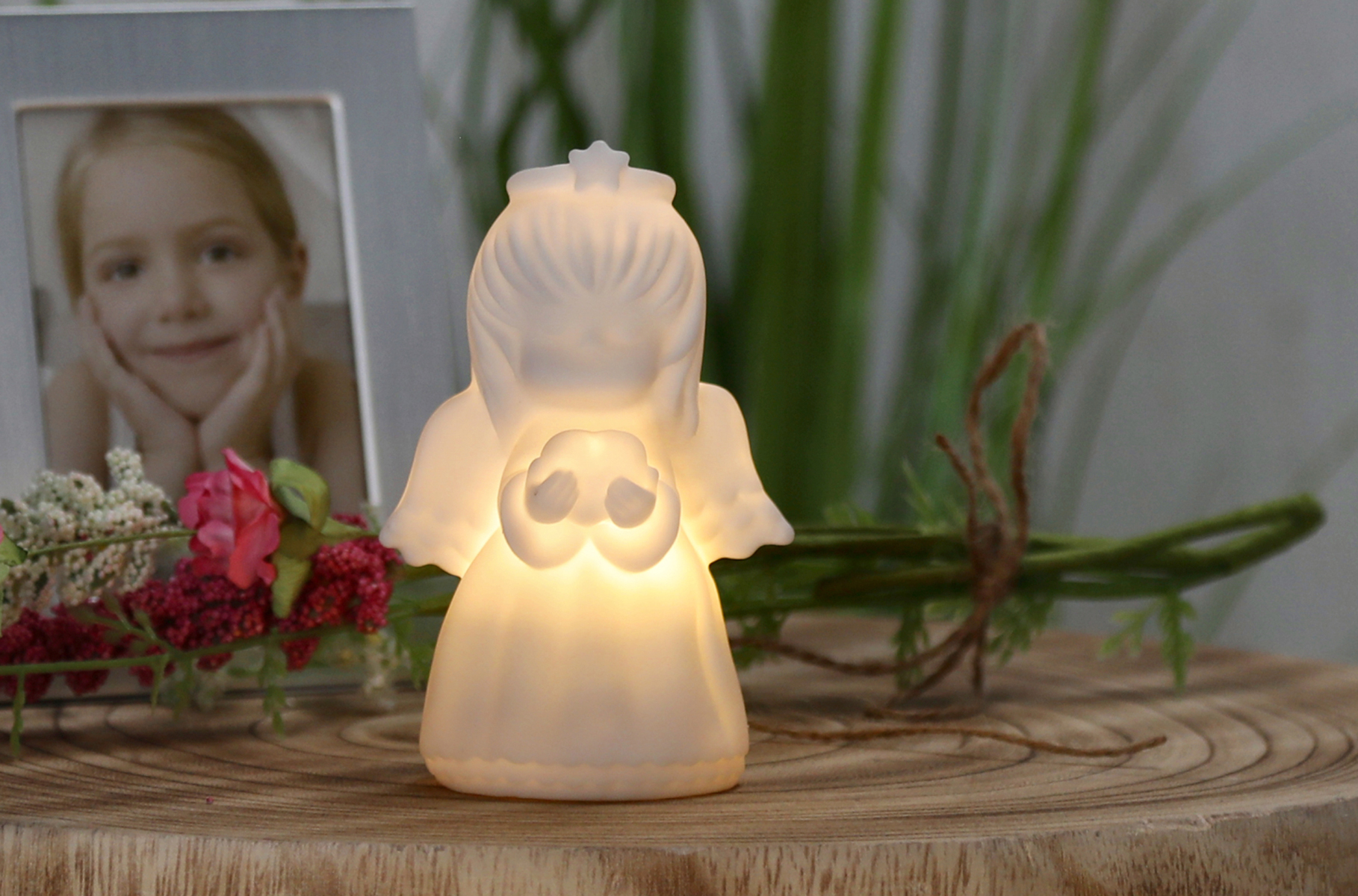 Leuchtende Engel-Figur aus Kunststoff. Hält zwei kleine Herzen. Sanfte, warme Lichtquelle. Steht auf einem Holzuntergrund, umgeben von Blumen und einem Bilderrahmen.