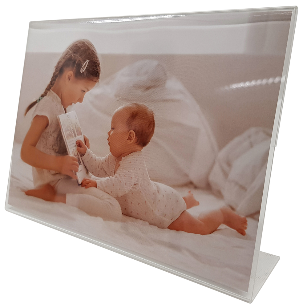 Ein Mädchen mit geflochtenem Haar sitzt auf einem Bett und hält ein Buch. Ein Baby liegt auf dem Bauch und schaut das Buch an. Helle, neutrale Farben, weiche Bettwäsche im Hintergrund.